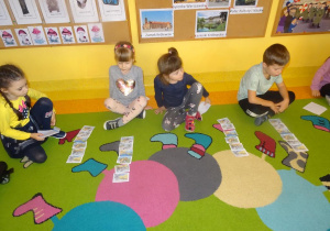 Czwórka dzieci siedzi na dywanie z rozłożonymi przed sobą obrazkami, układają historyjkę obrazkową.